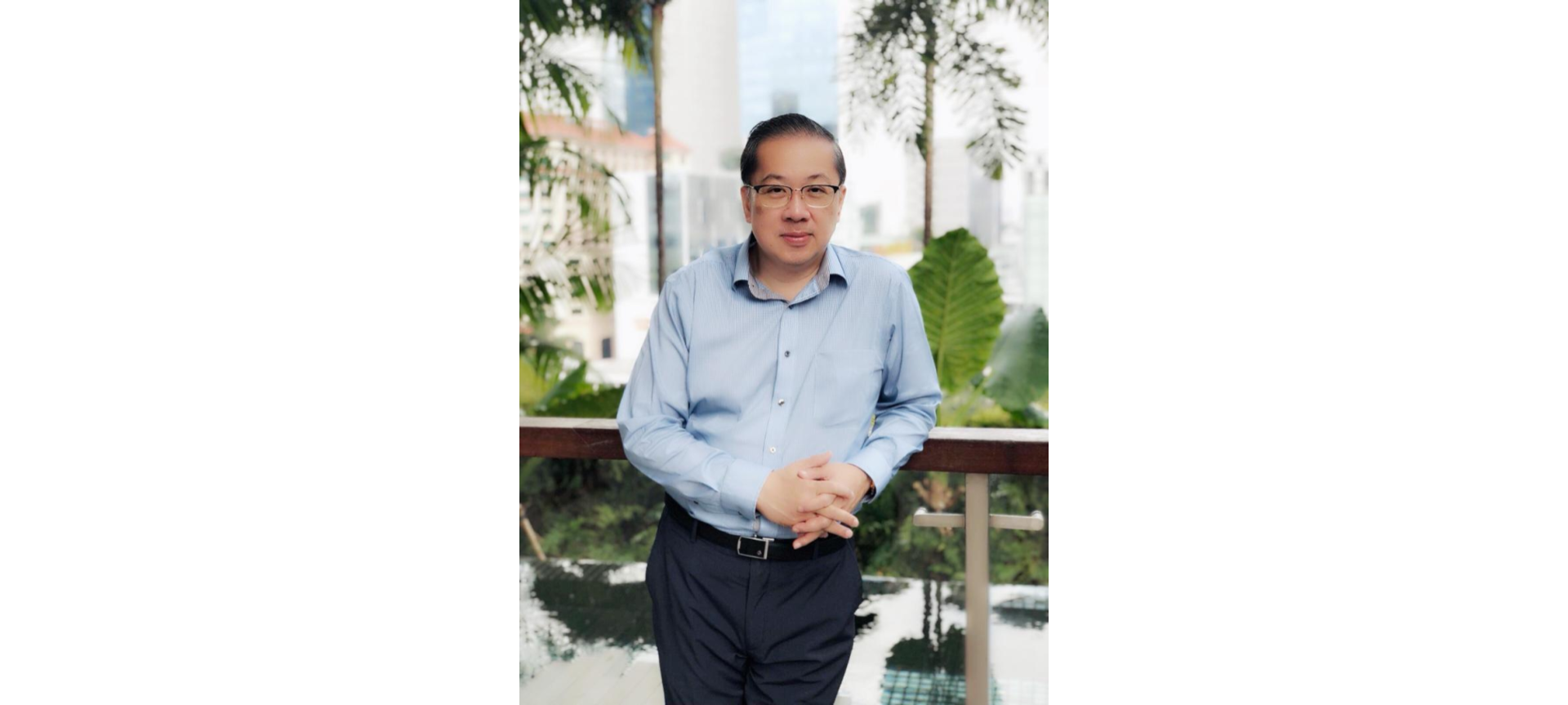 A man in a light blue dress shirt and black pants, standing with hands clasped in front of a balcony with greenery.